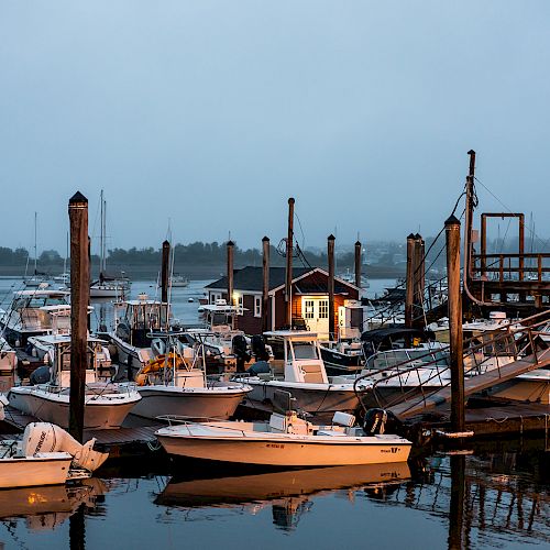 A serene marina scene at dusk, with multiple boats docked, calm waters, and a small illuminated building on the pier, creating a tranquil atmosphere.