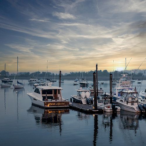 The image shows a serene marina at sunset with several boats docked and a calm water surface reflecting the sky and vessels.