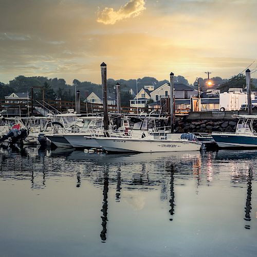 A peaceful harbor scene at sunset, with several boats docked by the pier and houses in the background, under a partly cloudy sky.