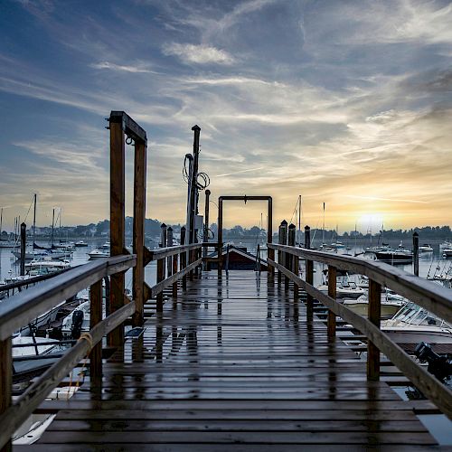 A wooden pier with railings leads to a marina filled with boats, under a picturesque sky with the sun setting or rising in the background.