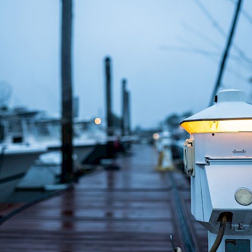 The image shows a dock with boats moored, and a lit lamp post in the foreground on an overcast day, creating a serene maritime scene.