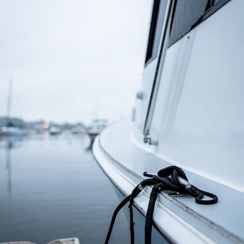 A close-up of a boat's side, featuring a tied rope, with a foggy marina and several boats faintly visible in the blurred background.