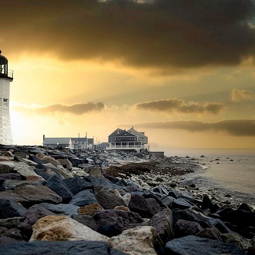 A scenic view of a lighthouse on a rocky shoreline at sunset, with buildings and a calm sea in the background, under a dramatic sky.