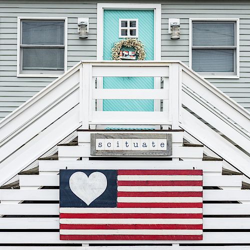 A gray house with a turquoise front door, white stairs, a wreath, an American flag with a heart, and a sign that reads 