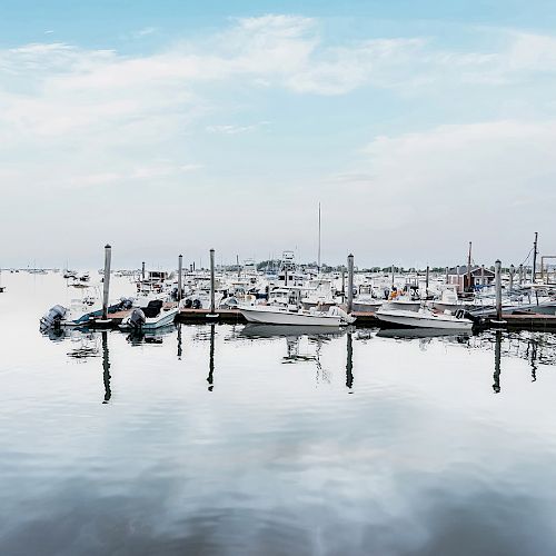 The image shows a calm marina with numerous boats docked at piers, surrounded by water and a cloudy sky. The water reflects the boats and the sky.