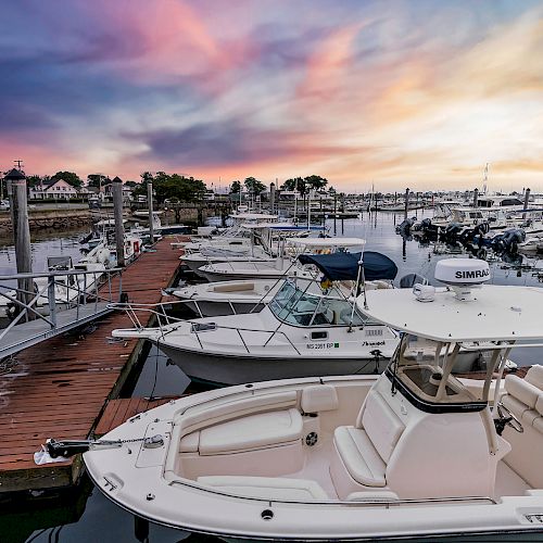 The image shows a marina at sunset, filled with various boats docked alongside wooden piers, with colorful skies in the background.