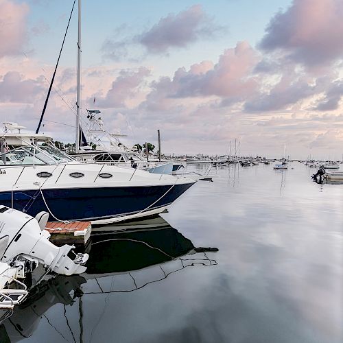 A serene harbor with boats docked on calm water, reflecting the soft pink and blue hues of the sky and scattered clouds.