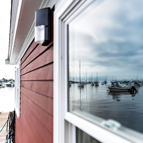 A red wooden building with white trim reflects a serene harbor filled with boats and sailboats under a cloudy sky, seen from a dock.