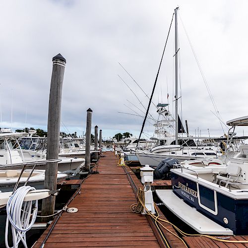 A wooden dock with boats and yachts on both sides is seen under a cloudy sky, with ropes and poles along the pathway.