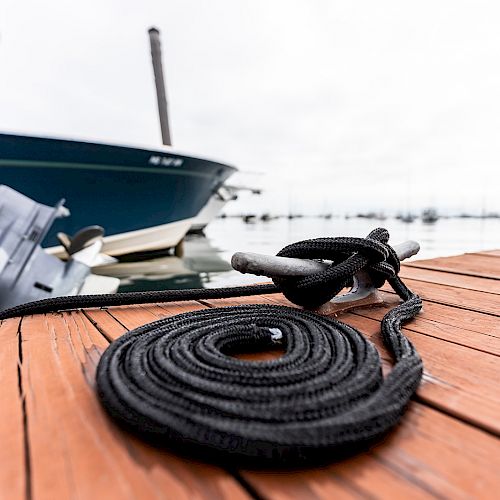 A coiled black rope is on a wooden dock, with a boat docked nearby and water in the background on a cloudy day.