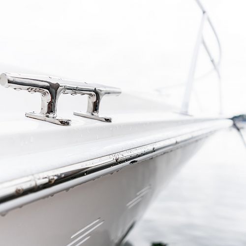 A close-up of a boat's deck showcasing a shiny metal cleat. The background is blurred, revealing a calm body of water and distant trees.