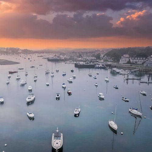 A serene harbor scene at sunset with numerous boats anchored in the water, surrounded by buildings and green landscape.