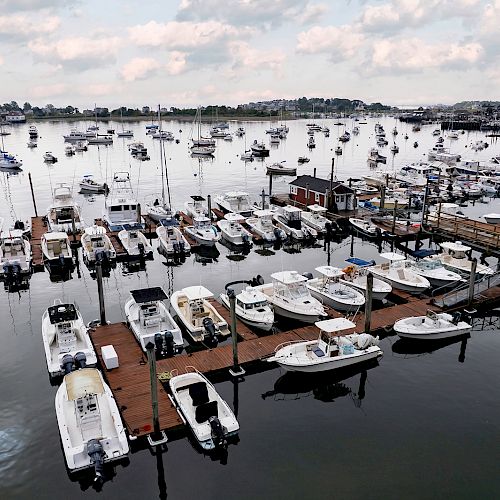 The image shows a marina with numerous boats docked at various piers, calm water, and a backdrop of more boats anchored further out.