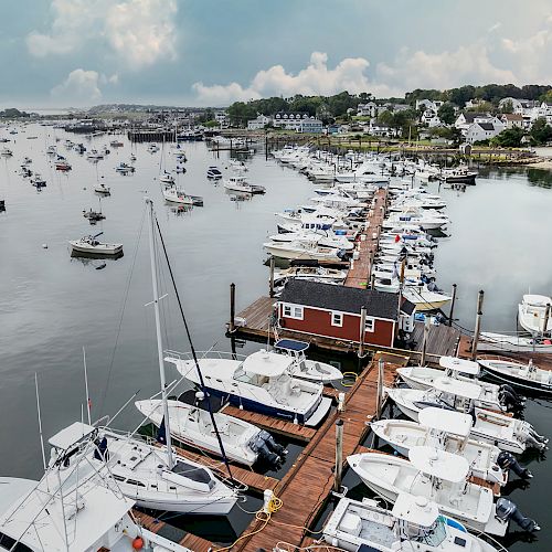 A scenic view of a marina with numerous boats docked along wooden piers, surrounded by calm water and coastal houses under a partly cloudy sky.