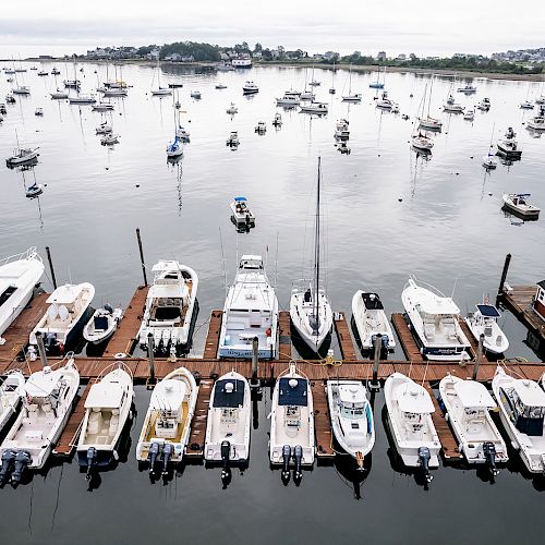 The image shows a dock with several boats moored, surrounded by a calm water harbor with more boats in the distance under an overcast sky.