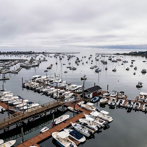 An aerial view of a marina with numerous boats docked at piers and sailboats anchored in the water, under a cloudy sky.