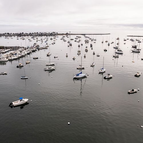 A calm harbor with numerous anchored boats and yachts, surrounded by a breakwater, and a distant shoreline on a cloudy day.