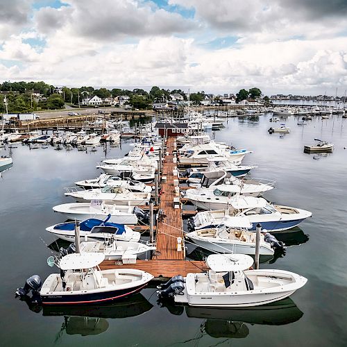 A marina with several boats docked along a wooden pier, surrounded by calm water and cloud-covered sky in the background.