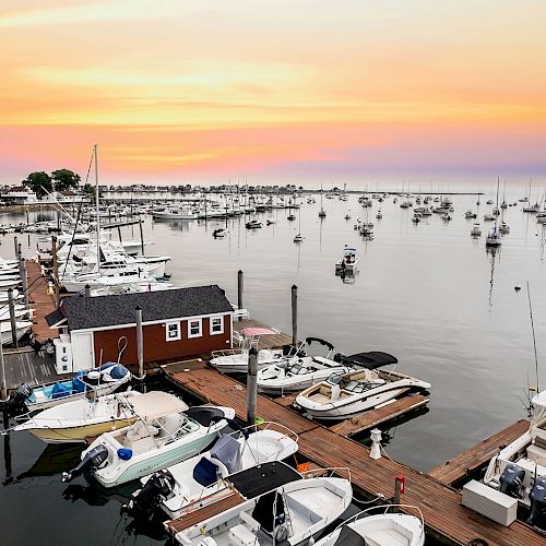 A serene harbor scene at sunset with boats docked at piers, a red boathouse, and numerous yachts anchored in the calm water.