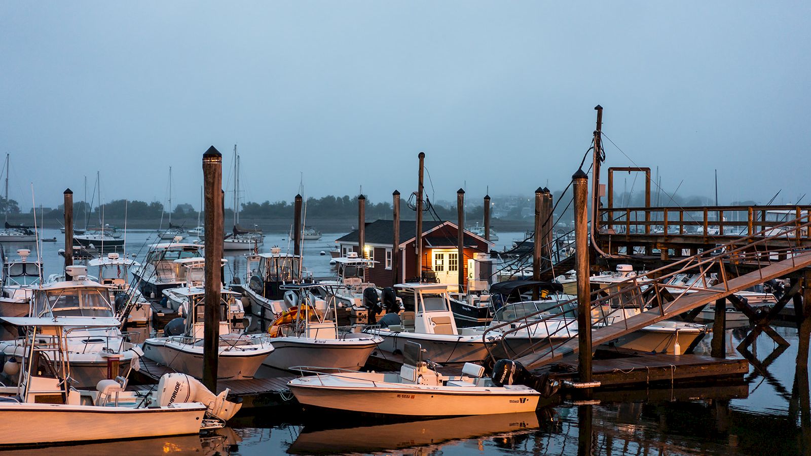The image depicts a marina with numerous boats docked, a small house nearby, and a wooden pier extending into the water.