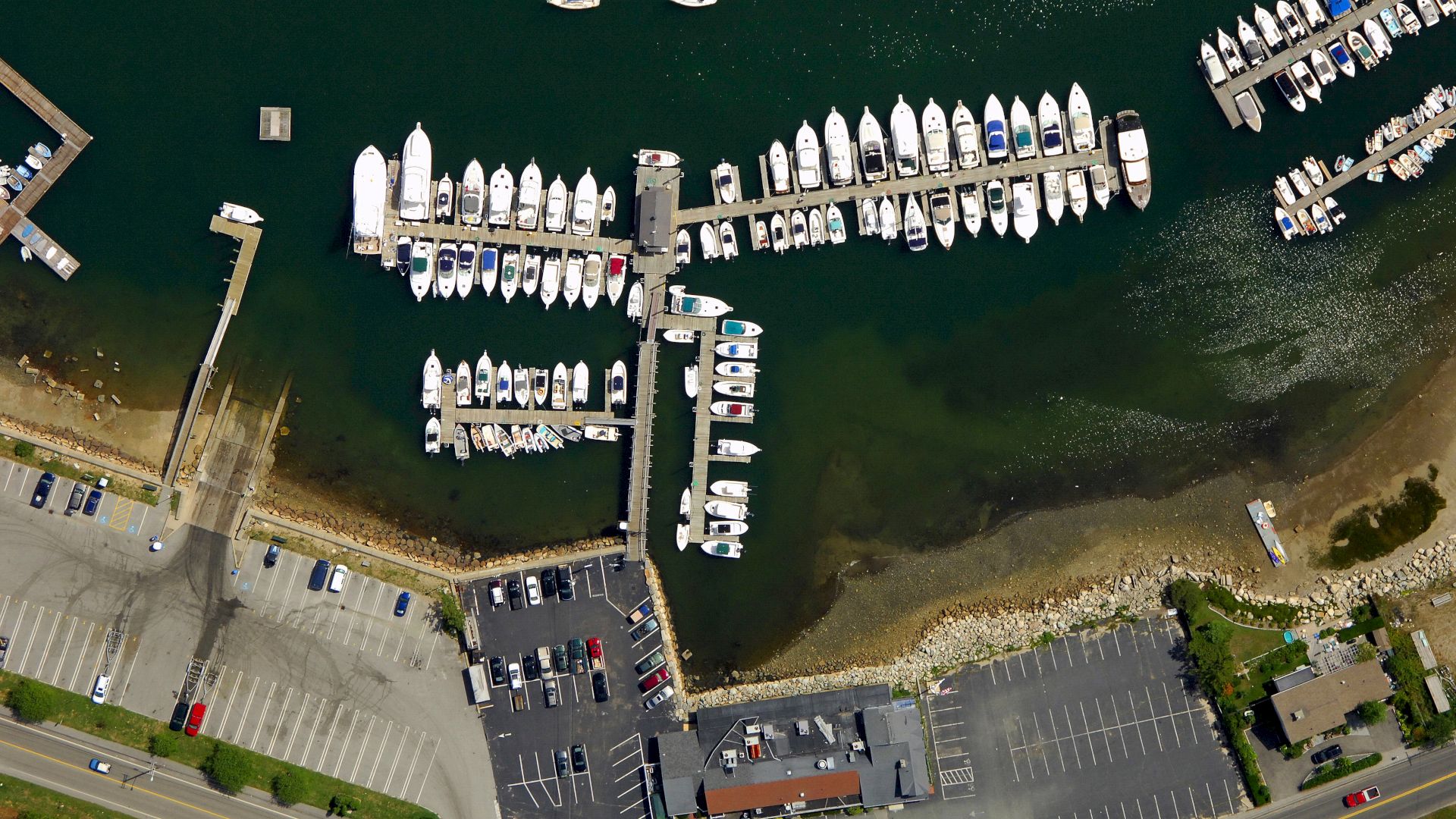 An aerial view of a marina showing boats docked, parking lots, roads, and nearby buildings along the shoreline, ending the sentence.