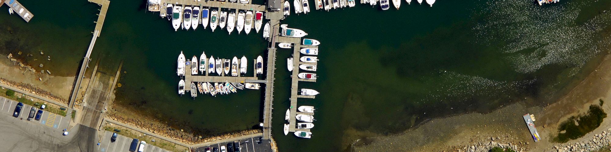 An aerial view of a marina showing boats docked, parking lots, roads, and nearby buildings along the shoreline, ending the sentence.